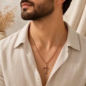 Puede incluir: Un colgante de cruz de metal en tono oro rosa suspendido de una cadena de eslabones a juego. La cruz presenta un acabado pulido con una ranura vertical central, sobre una camisa de botones texturizada de color claro.