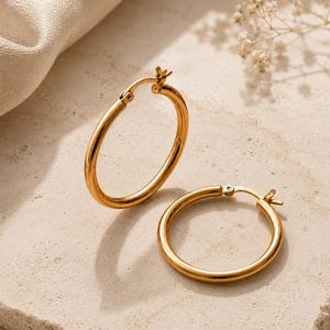 May include: A pair of gold hoop earrings with a polished finish. One earring stands upright while the other lies flat. Each hoop features a hinged post closure mechanism at the top, showcasing a simple, circular design made of smooth, reflective metal.
