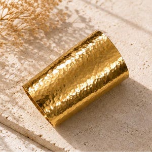 Può includere: Un ampio bracciale a fascia cilindrico realizzato in metallo lucido color oro. La superficie presenta una distinta texture martellata che crea numerose piccole rientranze riflettenti su tutto l'esterno del pezzo, conferendogli un aspetto metallico brillante.