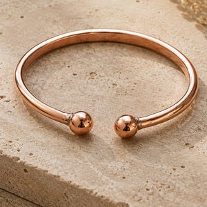 Peut inclure: Un bracelet ouvert de couleur or rose avec une finition lisse et polie. Le bracelet circulaire présente un design simple et épuré, se terminant à chaque extrémité par une petite boule sphérique qui offre un aspect propre et équilibré.