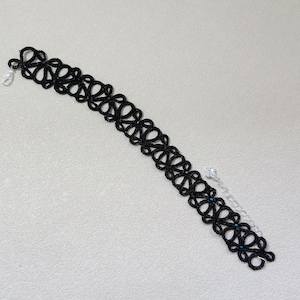 May include: A black lace choker necklace featuring a repeating floral pattern made of thin thread. The choker includes a silver-toned metal lobster claw clasp and a short silver-toned metal extender chain with small blue beads attached.