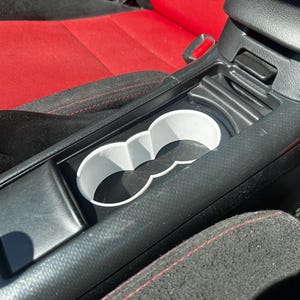 May include: A white plastic cup holder insert with a figure-eight shape, designed to fit into the center console of a vehicle. The insert features two circular openings and is positioned within the black plastic console between two red and black seats.