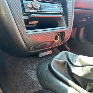 May include: A car interior center console featuring a carbon fiber patterned trim, a Pioneer head unit, a circular power outlet, and a small rectangular USB hub mounted below the console near the black leather gear shift boot.