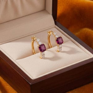 May include: A pair of gold hoop earrings featuring a central rectangular deep pink gemstone flanked by small clear stones. The earrings are presented in a dark wooden box with a cream-colored interior lining, designed for secure jewelry storage.