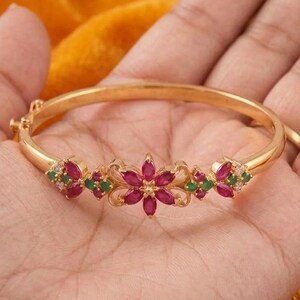 May include: A gold-toned bangle bracelet featuring a central flower motif set with marquise-cut magenta stones and small round green stones. The metal band is smooth and circular, with additional floral-inspired accents flanking the center piece.