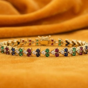 May include: A gold-toned bracelet featuring a repeating pattern of multi-colored round gemstones. The design includes alternating clusters of clear white, deep blue, vibrant green, and rich red stones set in a linked metal chain structure.
