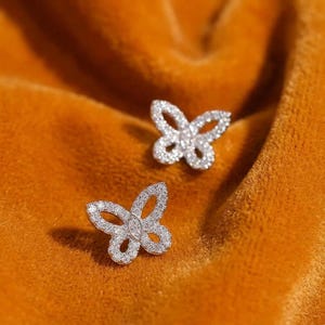 May include: A pair of silver-toned butterfly-shaped stud earrings, each featuring a delicate open-wing design encrusted with numerous small, clear, sparkling stones that cover the entire surface of the butterfly wings and body.