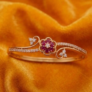 Puede incluir: Un brazalete de oro con un motivo floral central compuesto por seis rubíes de color rojo intenso que rodean un diamante blanco claro. La banda está acentuada con pequeñas piedras blancas claras y delicados detalles de filigrana de metal.