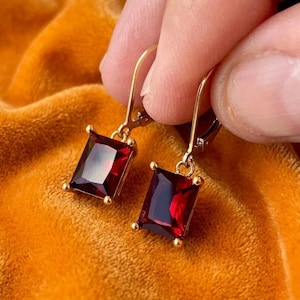 May include: A pair of gold-toned drop earrings featuring rectangular, faceted deep red gemstones. Each earring is suspended from a simple gold hook wire, showcasing a clear, polished finish on the stones that catch the light with a vibrant crimson hue.