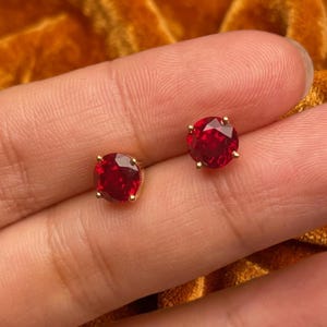 May include: A pair of round, faceted red gemstone stud earrings set in gold-toned metal prongs. Each earring features a circular, deep red stone with a brilliant cut that reflects light, secured by four small metal prongs on each side.