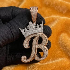 May include: A rose gold pendant in the shape of the letter B, featuring a script font encrusted with small clear stones. A crown design sits atop the letter, also covered in clear stones, with a rectangular bail at the very top for a chain.