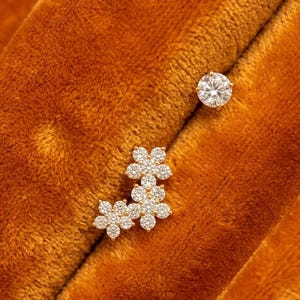 May include: A pair of gold earrings featuring one single round brilliant-cut clear stone and one cluster earring composed of three small flower-shaped arrangements, each set with multiple tiny clear stones that catch the light.