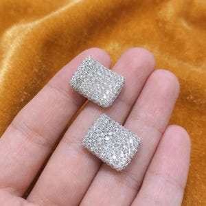 May include: A pair of rectangular stud earrings featuring a dense pavé setting of small, clear, reflective stones. The earrings have a metallic silver-toned base and a flat, uniform surface covered entirely in sparkling, light-catching crystals.