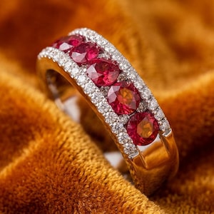 May include: A gold ring featuring a central row of five round-cut, deep red rubies. The rubies are flanked on both sides by two parallel rows of small, clear, round-cut diamonds set into the polished gold band, creating a multi-stone design.