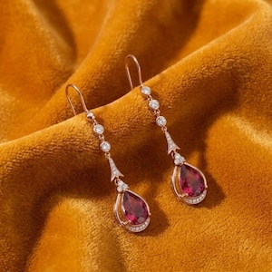 Puede incluir: Un par de pendientes colgantes de oro rosa con una piedra preciosa de color rojo intenso en forma de lágrima en la base, acentuados por una serie de pequeñas piedras redondas transparentes a lo largo de la barra metálica y la gema central.