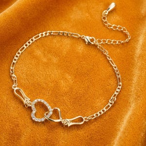May include: A gold-toned chain bracelet featuring a central design of two interlocking heart-shaped links, one of which is encrusted with small clear stones. The bracelet includes a delicate link chain and a small teardrop-shaped charm at the end.