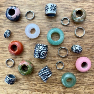 May include: Assortment of colorful gemstone beads and silver hair tie charms. The beads are round and have various patterns and colors, including green, pink, brown, and white. The charms are silver and have different designs, including flowers and swirls.