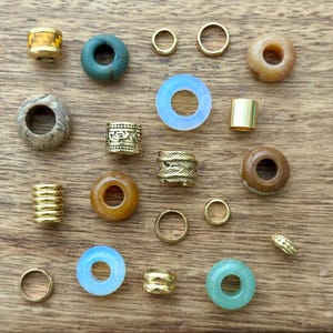 Puede incluir: Una colecci&oacute;n de cuentas y anillos de diversas formas y tama&ntilde;os. Las cuentas incluyen cilindros de metal dorado, cuentas de metal con dise&ntilde;os y cuentas de piedra en tonos marr&oacute;n, verde y azul. Tambi&eacute;n hay varios anillos dorados.