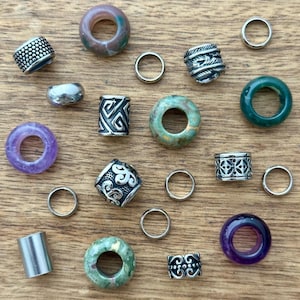 May include: An assortment of beads and rings in various colors and designs. The beads include silver, purple, green, and patterned styles. The rings are silver and vary in size. The items are arranged on a wooden surface.