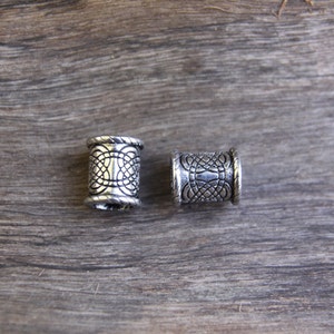 May include: Two silver metal hair beads with an intricate design. The beads are cylindrical and have a textured surface.