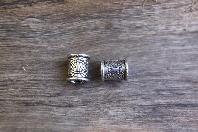 May include: Two silver metal hair beads with an intricate design. The beads are cylindrical and have a textured surface.