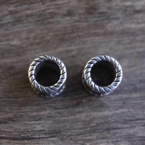 May include: Two silver ear gauges with a braided design. The gauges are round and have a hole in the center.