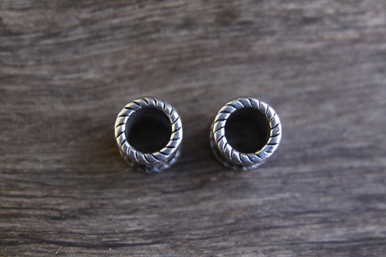 May include: Two silver ear gauges with a braided design. The gauges are round and have a hole in the center.
