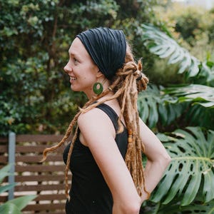 Peut inclure: Un bandeau noir et un débardeur assorti. La personne porte des boucles d'oreilles vertes et a de longues dreadlocks. L'arrière-plan comprend un feuillage vert et une clôture en bois.