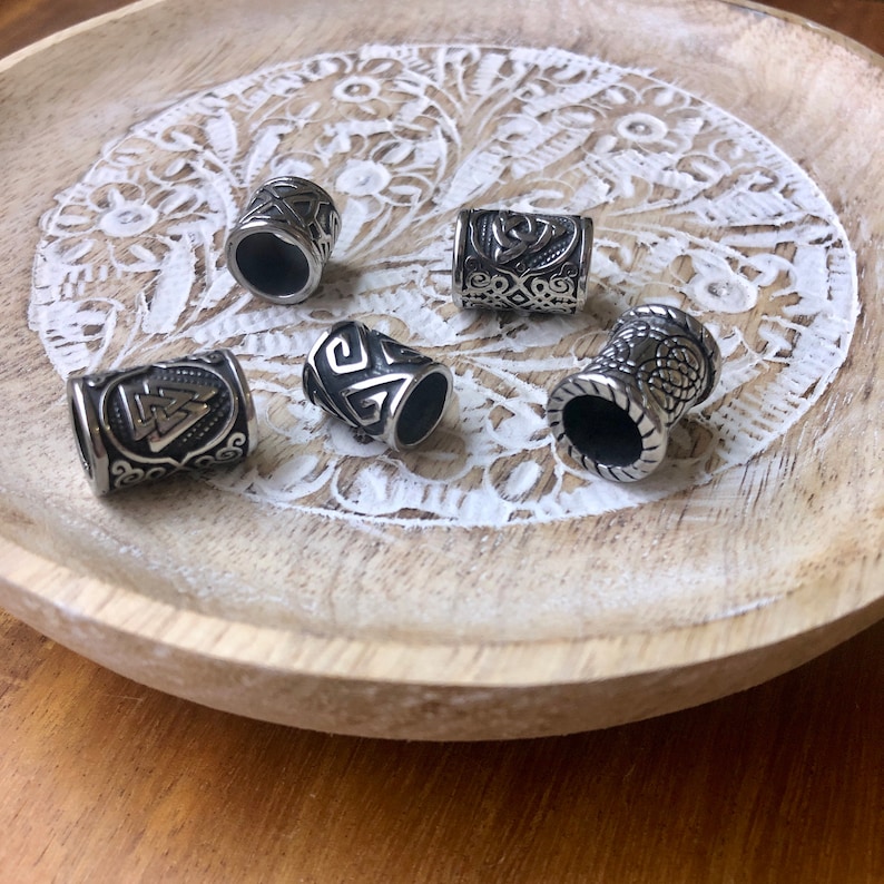 May include: Five silver metal hair beads with different designs, including a Celtic knot, a triskelion, and a geometric pattern. The beads are arranged on a white wooden surface.
