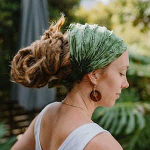Op de afbeelding: Een vrouw met een groene hoofddoek en grote bruine oorringen. De hoofddoek is gemaakt van een gekreukelde stof. De vrouw heeft lange, bruine dreadlocks en draagt een witte tanktop. Een gouden ketting is zichtbaar.