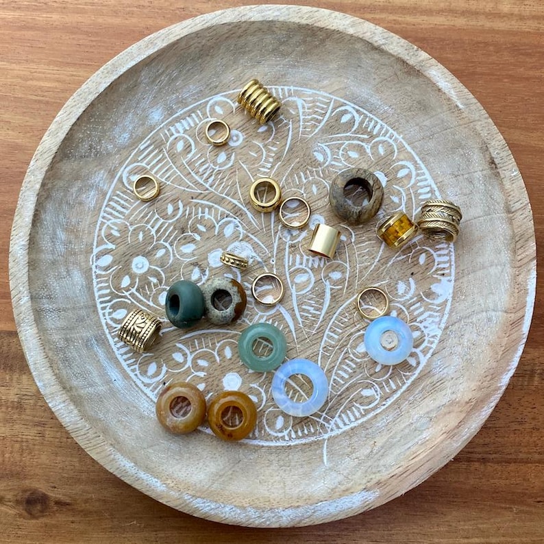 Puede incluir: Una colecci&oacute;n de cuentas y anillos de varios tama&ntilde;os y materiales, incluyendo metal dorado, madera y vidrio transl&uacute;cido. Las cuentas est&aacute;n dispuestas en una bandeja redonda de madera clara con un dise&ntilde;o floral blanco tallado.