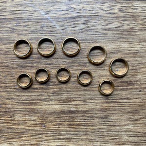 Op de afbeelding: Een verzameling van tien goudkleurige ringen van verschillende maten, gerangschikt op een houten oppervlak. De ringen zijn cirkelvormig met een gladde, gepolijste afwerking en zijn gelijkmatig verdeeld.