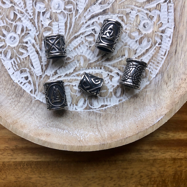 May include: Five silver metal hair beads with different Norse symbols engraved on them. The beads are arranged on a white wooden surface with a carved pattern.