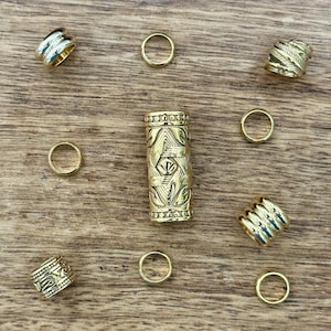 Puede incluir: Cuentas y anillos para el cabello de color dorado dispuestos sobre una superficie de madera. La cuenta central es cilíndrica con un intrincado diseño grabado. Varios anillos más pequeños y otras cuentas decorativas con patrones texturizados están dispersos.