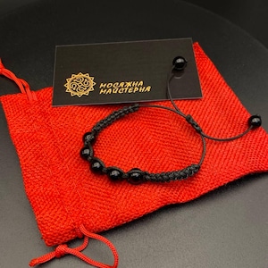 Könnte beinhalten: Ein schwarzes Kordelarmband mit glatten, runden schwarzen Perlen und einem geflochtenen Design. Das Armband liegt auf einem leuchtend roten Stoffbeutel neben einer schwarzen Karte mit goldenem Sonnenlogo und dem Text "МОСЯЖНА МАЙСТЕРНЯ".