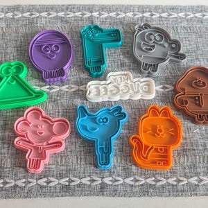 May include: A collection of nine plastic cookie cutters in various colours including green, purple, teal, grey, pink, blue, orange, and brown. One white cookie cutter features the text Hey Duggee. The shapes represent cartoon characters and objects.