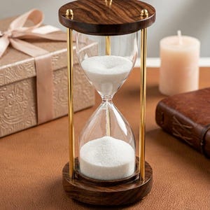 May include: An hourglass featuring a clear glass bulb filled with white sand, held between two circular dark wood bases. The structure is supported by three slender, polished gold-tone metal rods, with small gold-tone decorative knobs on the top base.