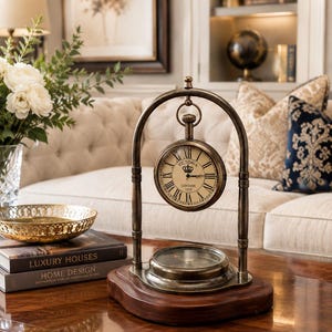 May include: A brass-toned metal desk clock suspended from an arched frame, mounted on a dark wooden base. The clock face features Roman numerals, a crown icon, and the text "VICTORIA LONDON 1915". A compass is integrated into the base below the clock.