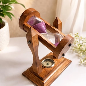 Puede incluir: Un marco de madera giratorio para reloj de arena con un bulbo de vidrio transparente que contiene arena morada. El marco está montado sobre una base rectangular de madera que presenta una brújula metálica circular incrustada en el centro.
