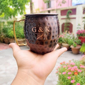 Puede incluir: Una taza de metal martillado color cobre con un asa de tono dorado pulido. El frente de la taza presenta el texto grabado G & K sobre un diseño de marcas de conteo. La superficie tiene un acabado texturizado con hoyuelos en todo el exterior.