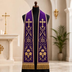 Könnte beinhalten: Eine violette liturgische Stola mit aufwendiger Goldstickerei. Das Design umfasst ein Kreuz oben, das IHS-Monogramm in der Mitte und Weinrebenmotive unten. Die Stola ist an den unteren Kanten mit goldenen Fransen versehen.
