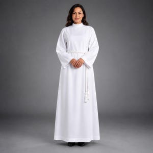 May include: A floor-length white alb featuring long sleeves, a high neckline, and a simple structure. A white rope cincture with two tassels is tied around the waist, cinching the fabric of the garment to create a clean, uniform appearance.