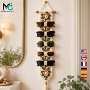 Puede incluir: Un tapiz vertical de ganchillo en color crema con un patrón de enredadera floral, hojas verdes y flores rosas. La pieza incluye cinco bucles integrados diseñados para sostener gafas de sol, suspendida de un soporte de madera con un cordón.
