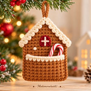Può includere: Una decorazione all'uncinetto a forma di casetta di pan di zenzero marrone con bordo del tetto color crema, una piccola finestra rossa ad arco con una croce bianca e un bastoncino di zucchero a righe rosse e bianche in una tasca color crema.