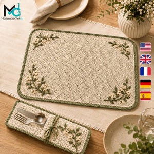 May include: A rectangular beige crochet placemat and a matching rectangular cutlery holder, both featuring a green border and embroidered floral sprigs in cream and green. The cutlery holder contains a silver fork and a silver spoon.