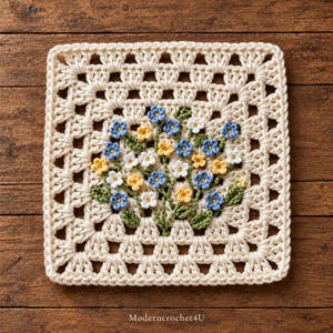 Puede incluir: Un cuadrado de ganchillo de color crema con un ramo central de pequeñas flores azules, amarillas y blancas con hojas verdes. El texto Moderncrochet4U está impreso en blanco en la parte inferior central.
