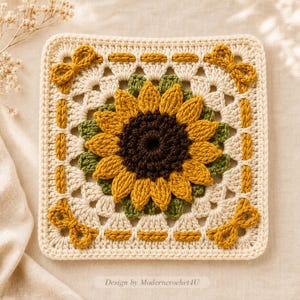 May include: A square crochet granny square featuring a central three-dimensional sunflower with a dark brown center and yellow petals, surrounded by green leaves. The border is cream with mustard yellow bows at each corner and text at the bottom.
