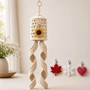 Puede incluir: Un adorno colgante cilíndrico de ganchillo color crema con un adorno de girasol y dos largas colas en espiral. Se incluyen tres pequeños dijes de ganchillo adicionales: una hoja de arce roja, un copo de nieve blanco y un corazón rosa.