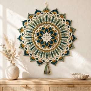 Könnte beinhalten: Ein kreisförmiger Wandbehang aus Häkelarbeit mit einem geschichteten Mandala-Design in den Farben Creme, Petrol, Salbeigrün und Senfgelb. Das komplexe Muster umfasst blütenblattartige Motive, offene Maschen und eine einzelne Quaste.