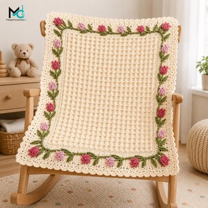 May include: A cream-colored crochet blanket featuring a waffle stitch center and a decorative border of green stems with pink and light pink tulip flowers. The blanket has a scalloped edge and is draped over a wooden rocking chair.